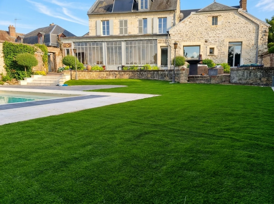 Jardin paysager avec pelouse synthétique vert vif, parfaitement tondue, bordant une piscine à débordement avec margelles en pierre blanche et plage en composite gris
